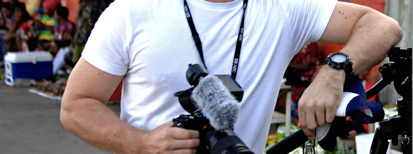 A young male journalist in a white tshirt with a camera around his neck leans against a tripod in a village street.